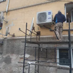 Instalator na rusztowaniu montuje jednostkę zewnętrzną klimatyzatora Haier na elewacji budynku z widocznymi śladami napraw i instalacjami satelitarnymi.