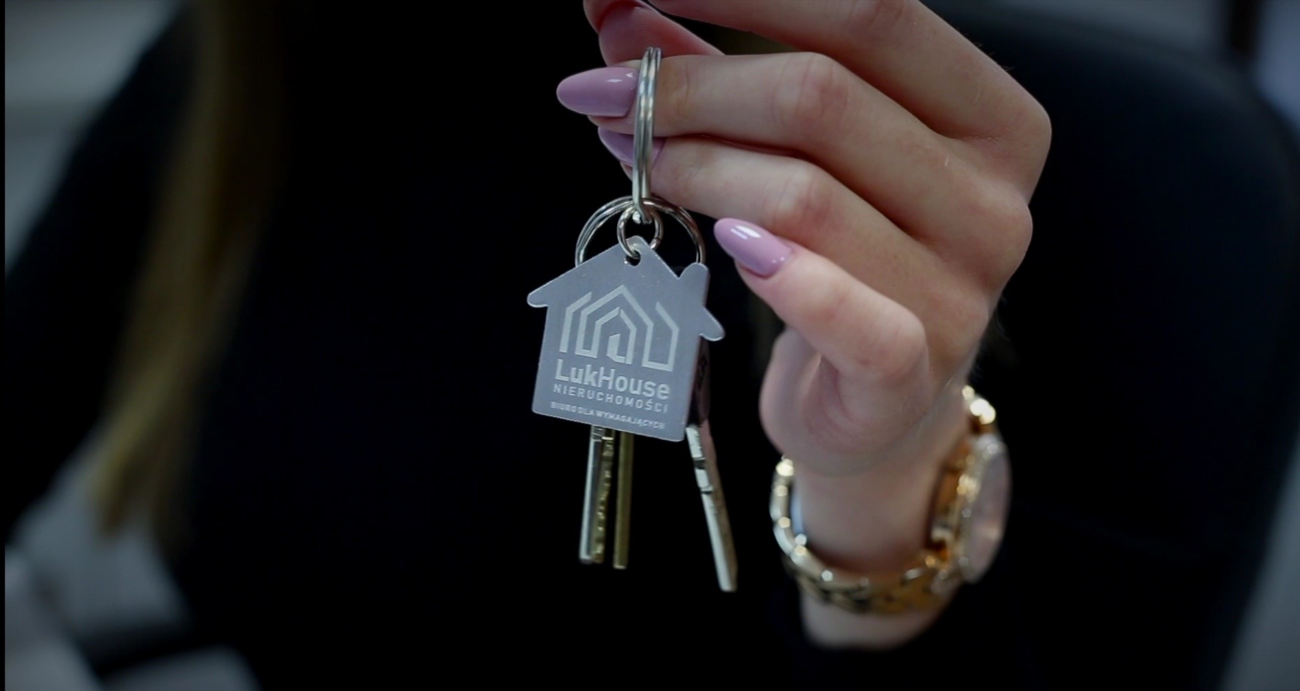 Klucze do domu z brelokiem LukHouse w dłoni kobiety z zegarkiem. Symbol zakupu lub wynajmu nieruchomości, pośrednictwo agencji.