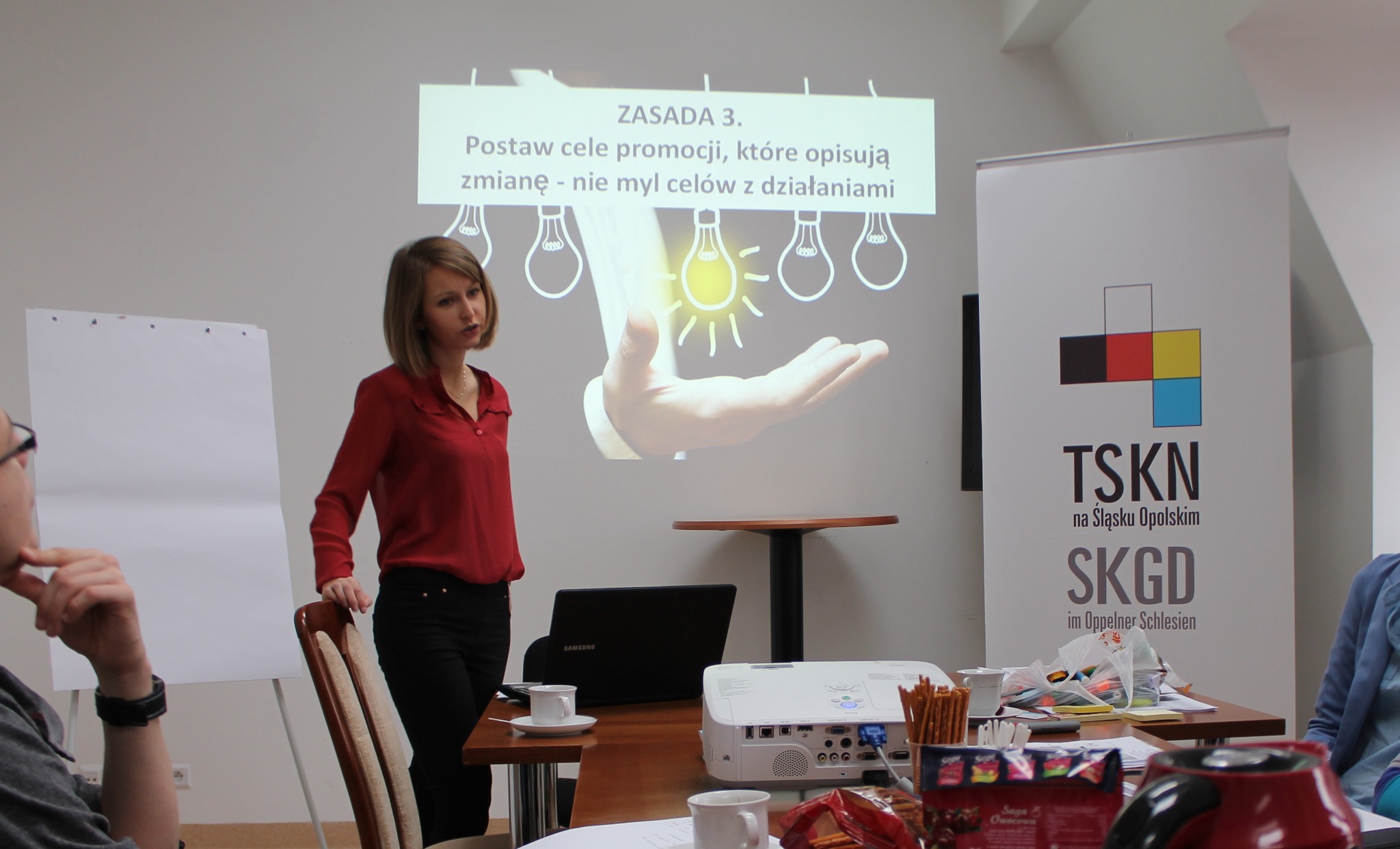 Prezentacja z zasadami promocji wyświetlana na ścianie, kobieta w czerwonej bluzce obok, laptop i projektor na stole.