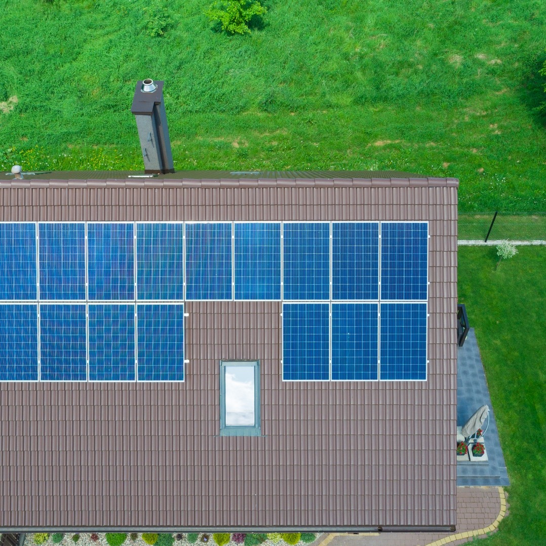 Widok z góry na brązowy dach z panelami słonecznymi w układzie dwurzędowym, kominem i oknem dachowym, otoczony zielonym trawnikiem i roślinami.