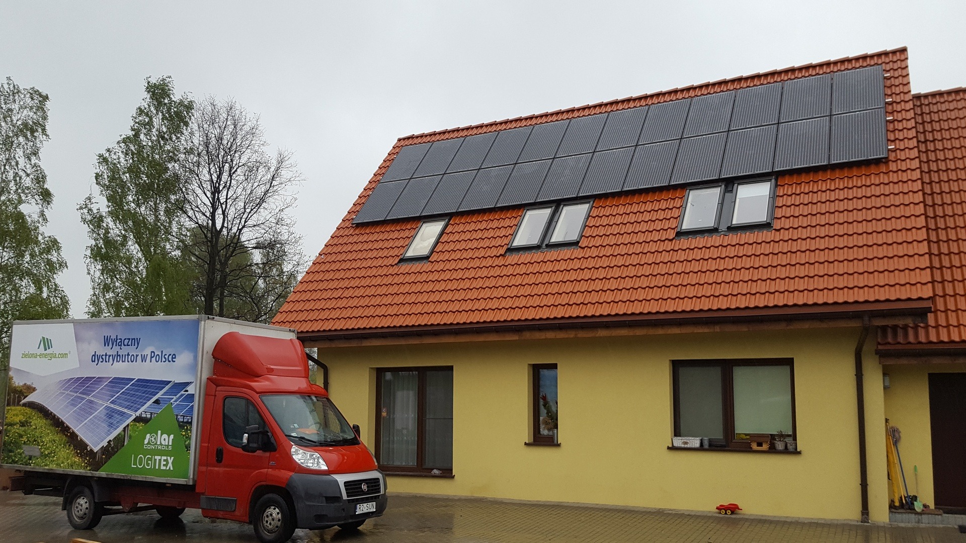 Czerwony samochód dostawczy z reklamą paneli słonecznych zaparkowany przed domem z zamontowanymi panelami fotowoltaicznymi na dachu pokrytym czerwoną dachówką.