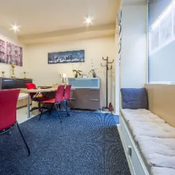 Jasne biuro z biurkiem, czerwonymi krzesłami, regałem z trofeami i kanapą przy oknie z widokiem na zimowe drzewa.