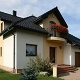 Dwukondygnacyjny dom jednorodzinny z beżowo-białą elewacją, czarnym dachem i balkonem z czerwonymi kwiatami w skrzynkach, widok z poziomu ogrodu.