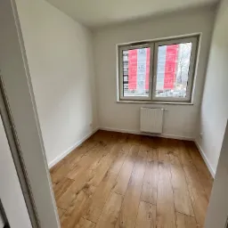 Pusty pokój z panelami podłogowymi w odcieniach brązu, białymi ścianami, grzejnikiem pod oknem z widokiem na budynek w rusztowaniach zabezpieczonych folią z logo.