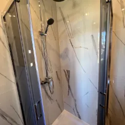 Otwarta kabina prysznicowa z chromowaną armaturą i marmurowymi płytkami na ścianach, widoczny odpływ liniowy w brodziku.