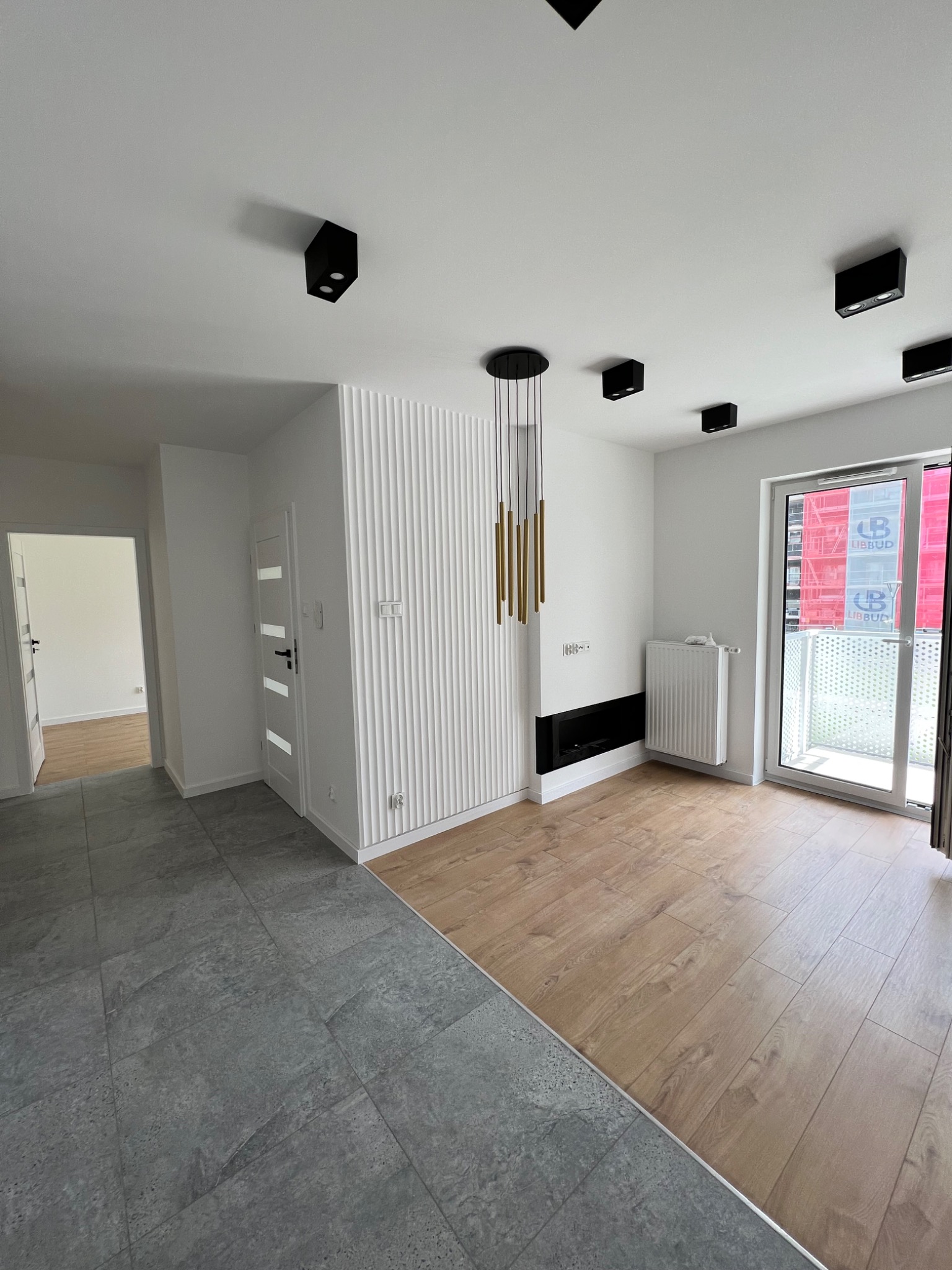 Nowoczesne wnętrze po remoncie: szara, kamienna podłoga przechodzi w drewnopodobne panele, białe ściany z dekoracyjnymi lamelami, minimalistyczne oświetlenie sufitowe i złota lampa wisząca...