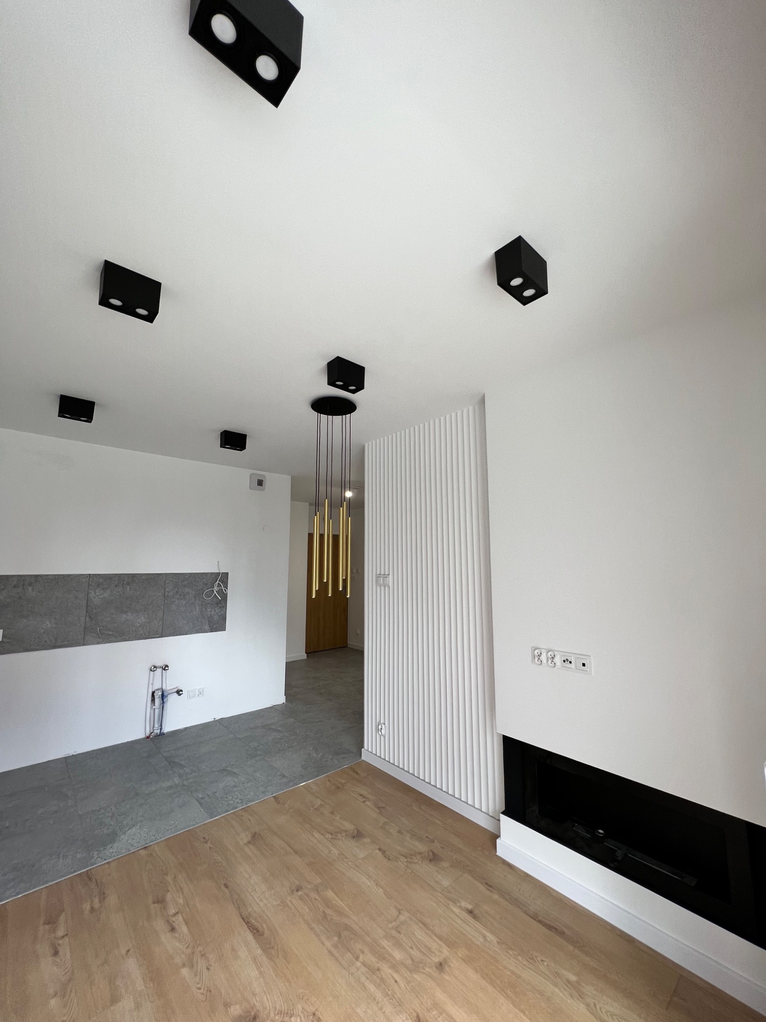 Surowe wnętrze w trakcie prac wykończeniowych: widoczne przyłącza wodne, szara ściana w miejscu przyszłej kuchni, panele podłogowe, dekoracyjna ściana z pionowymi żłobieniami i czarny wkład kominkowy.