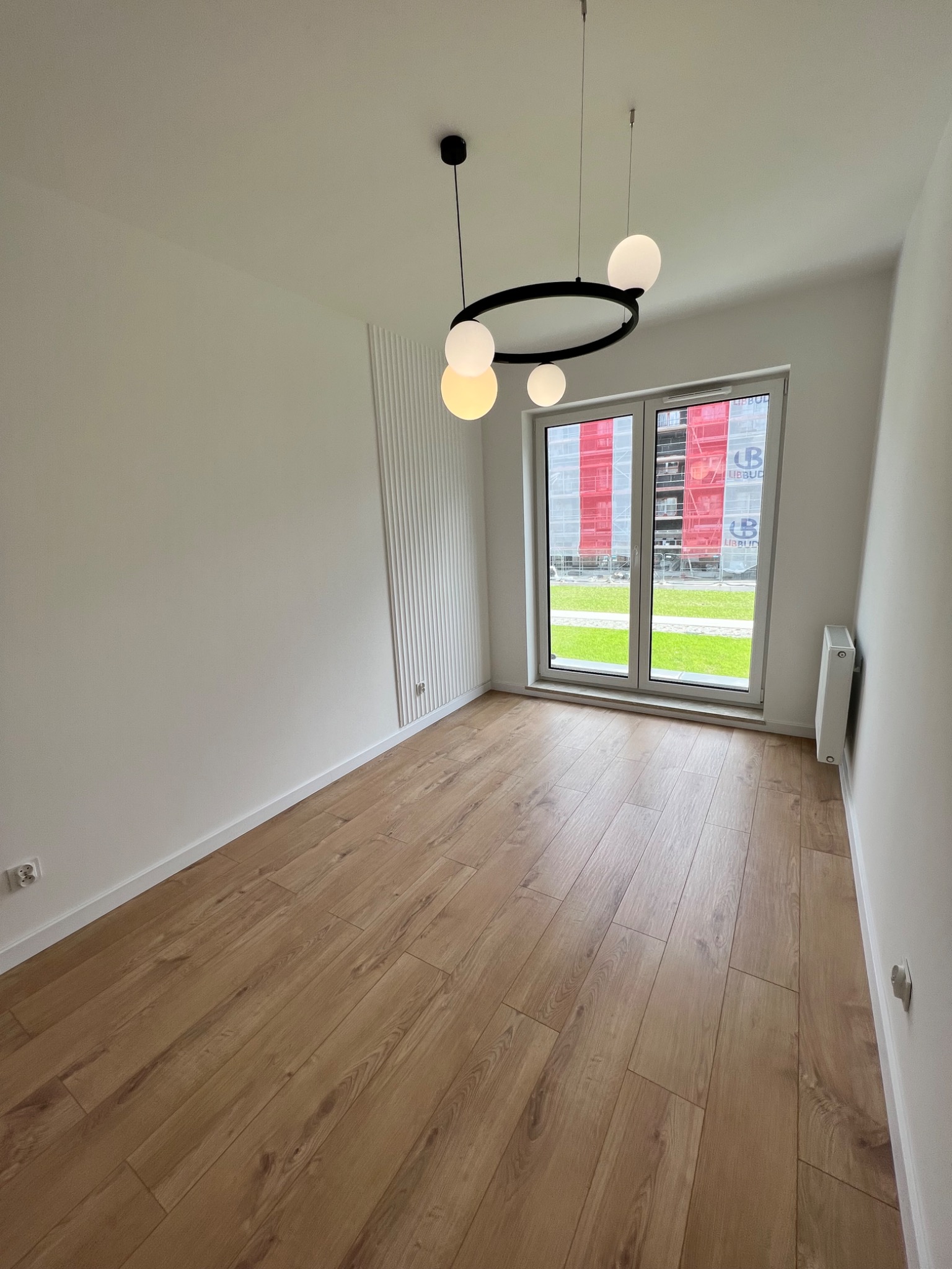 Pusty pokój z panelami podłogowymi w odcieniach drewna, białymi ścianami z dekoracyjnym panelem pionowym i nowoczesną lampą pierścieniową z kulistymi kloszami, okno z widokiem na budowę.