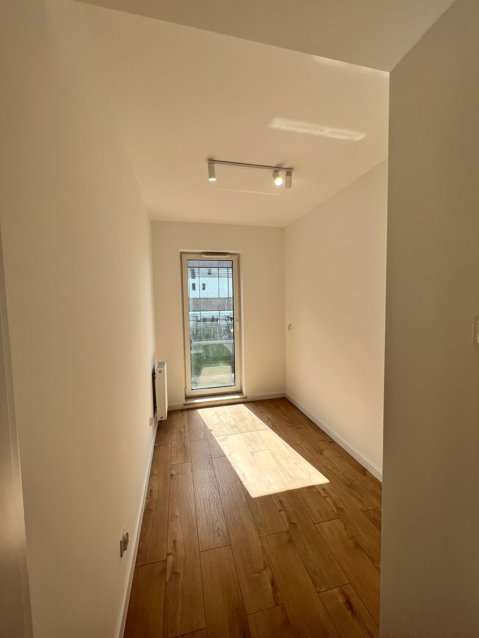 Pusty pokój po remoncie z drewnianą podłogą, białymi ścianami, oświetleniem punktowym na suficie i oknem balkonowym wpuszczającym naturalne światło.