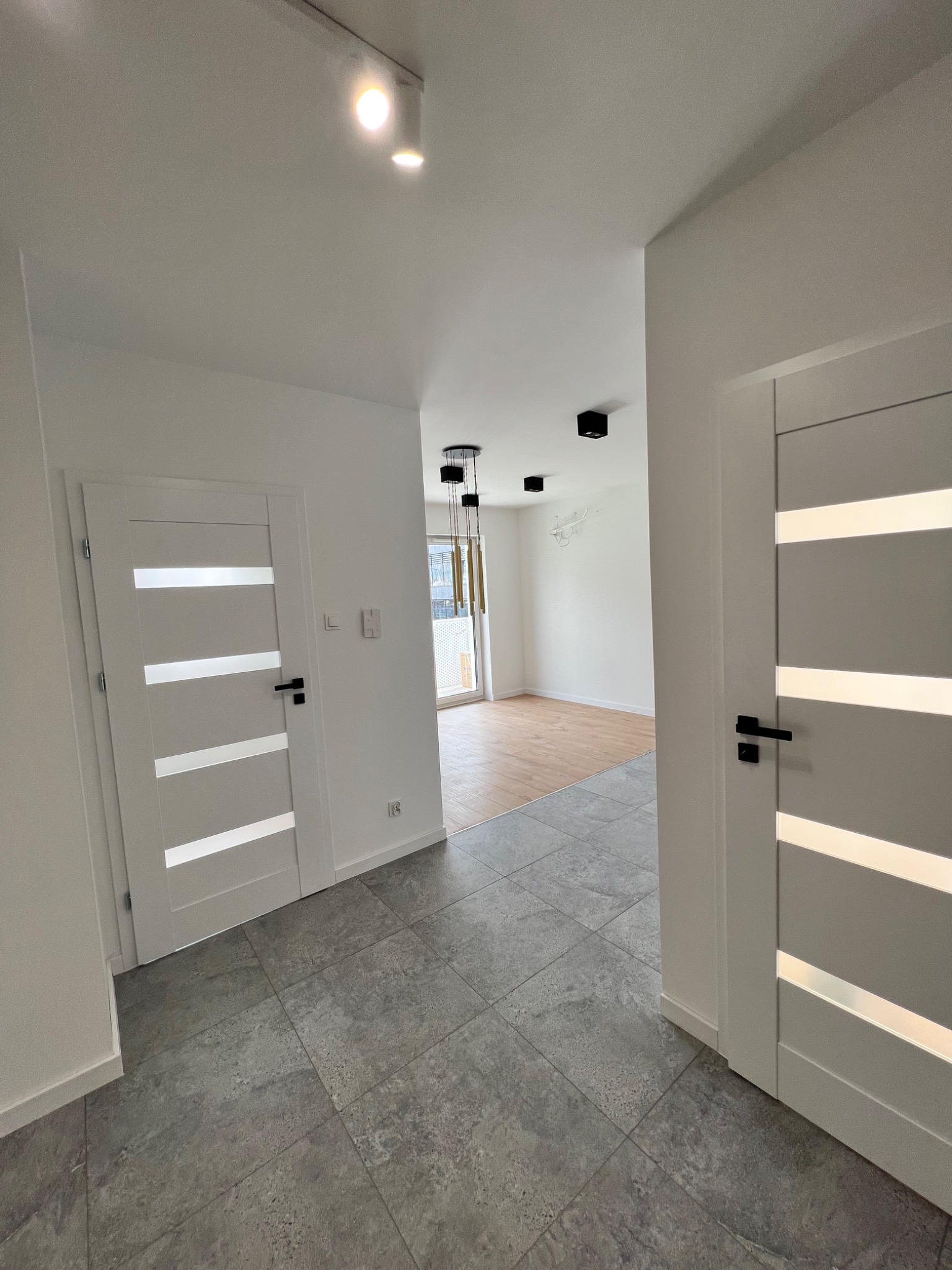 Nowoczesny korytarz z szarą podłogą z płytek, białymi ścianami i drzwiami z przeszkleniami, widok na salon z drewnianą podłogą i minimalistycznym oświetleniem.