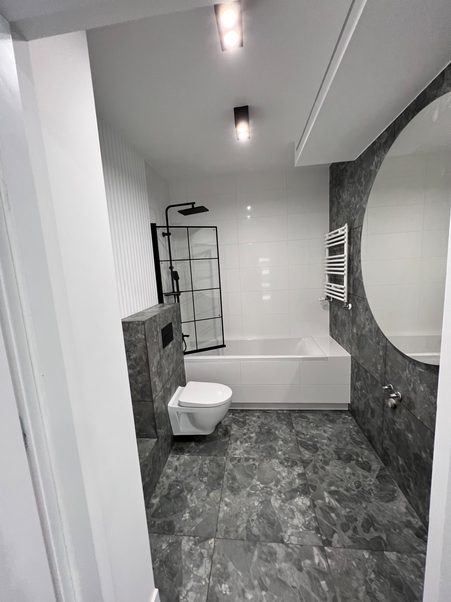 Nowoczesna łazienka z szarymi marmurkowymi płytkami na podłodze i ścianie, białą wanną, wiszącą toaletą i czarną kabiną prysznicową typu walk-in.
