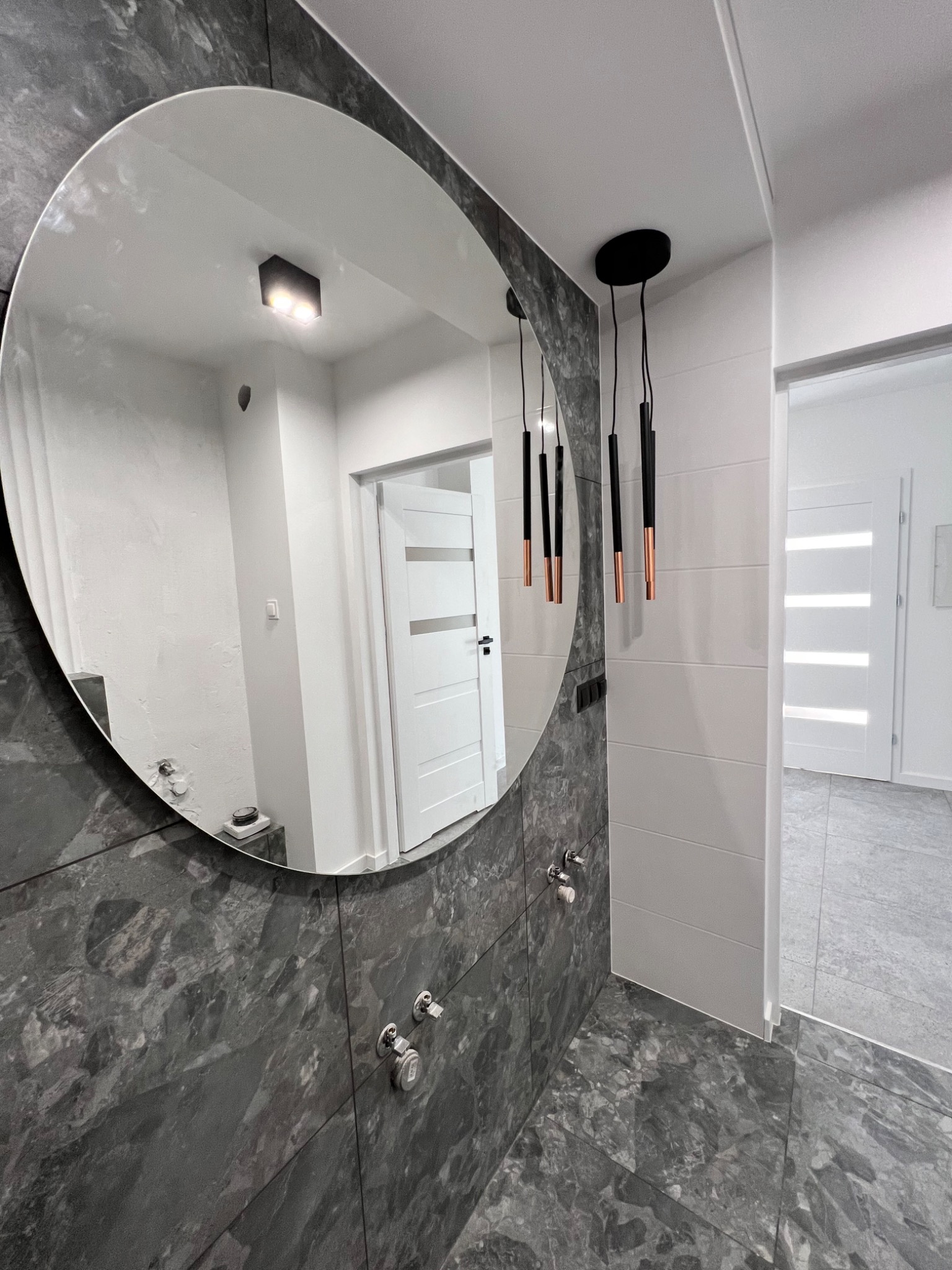 Nowoczesna łazienka z owalnym lustrem na ścianie z płytek imitujących marmur, wiszące lampy z czarnymi i miedzianymi elementami, oraz widok na korytarz z białą ścianą i drzwiami.