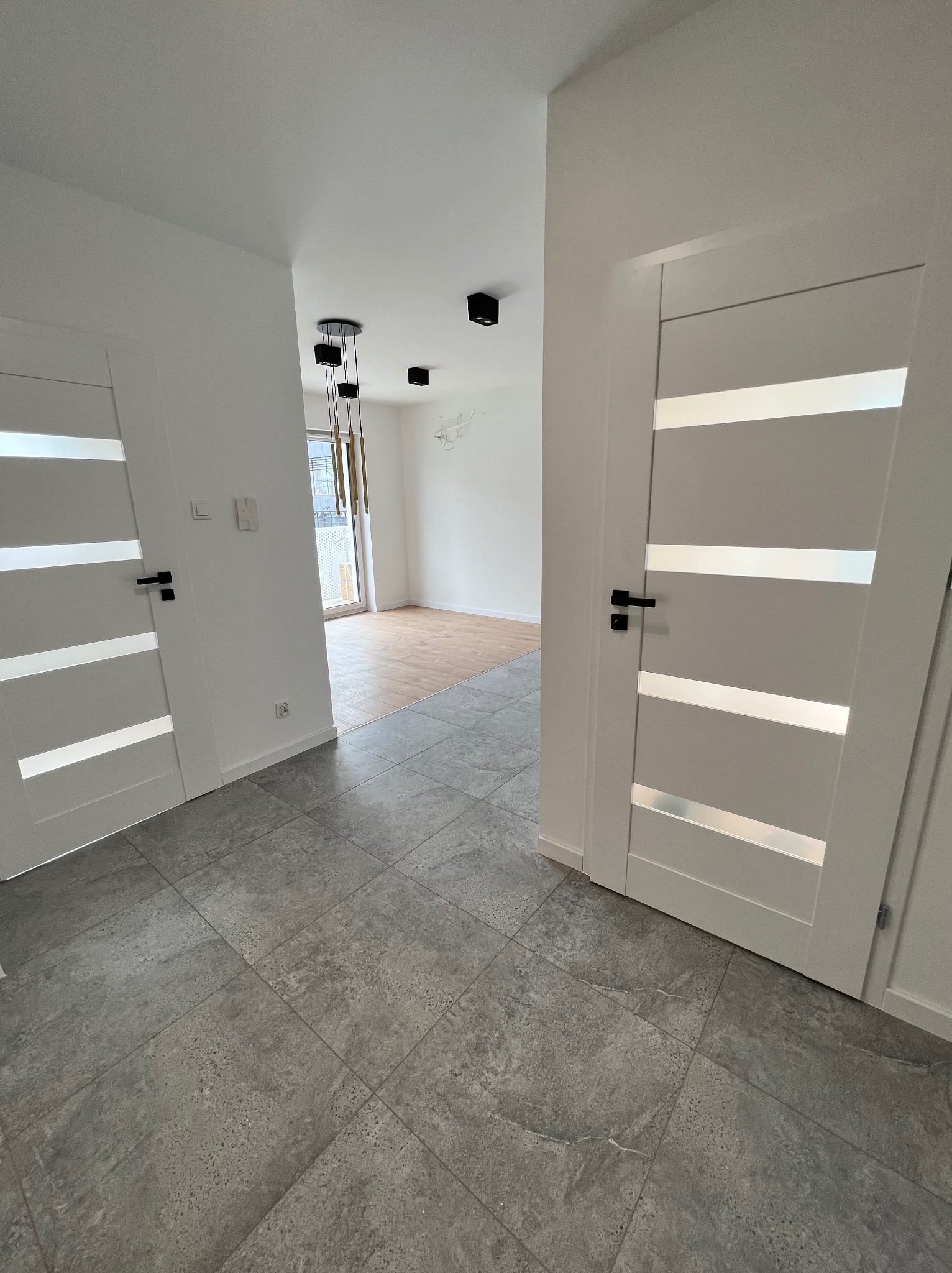 Wnętrze mieszkania po remoncie: szara, kwadratowa podłoga z płytek, białe drzwi z poziomymi, przezroczystymi paskami, drewniana podłoga w oddali, czarne lampy sufitowe.