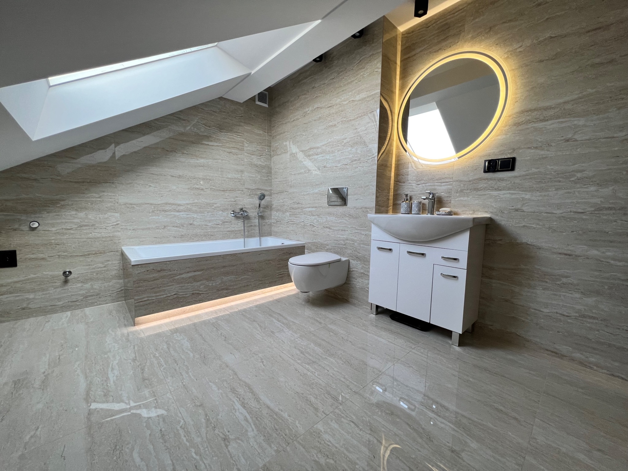Jasna łazienka na poddaszu z oknem dachowym, wykończona płytkami imitującymi marmur, z podświetlaną wanną, wiszącą toaletą i okrągłym lustrem z podświetleniem LED.