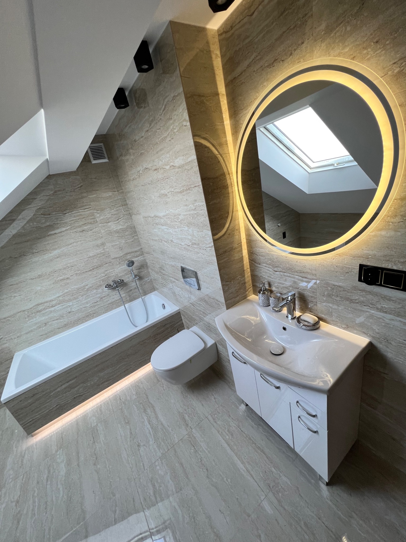 Nowoczesna łazienka na poddaszu z beżowymi płytkami imitującymi marmur, oświetlonym lustrem, białą armaturą i oknem dachowym.