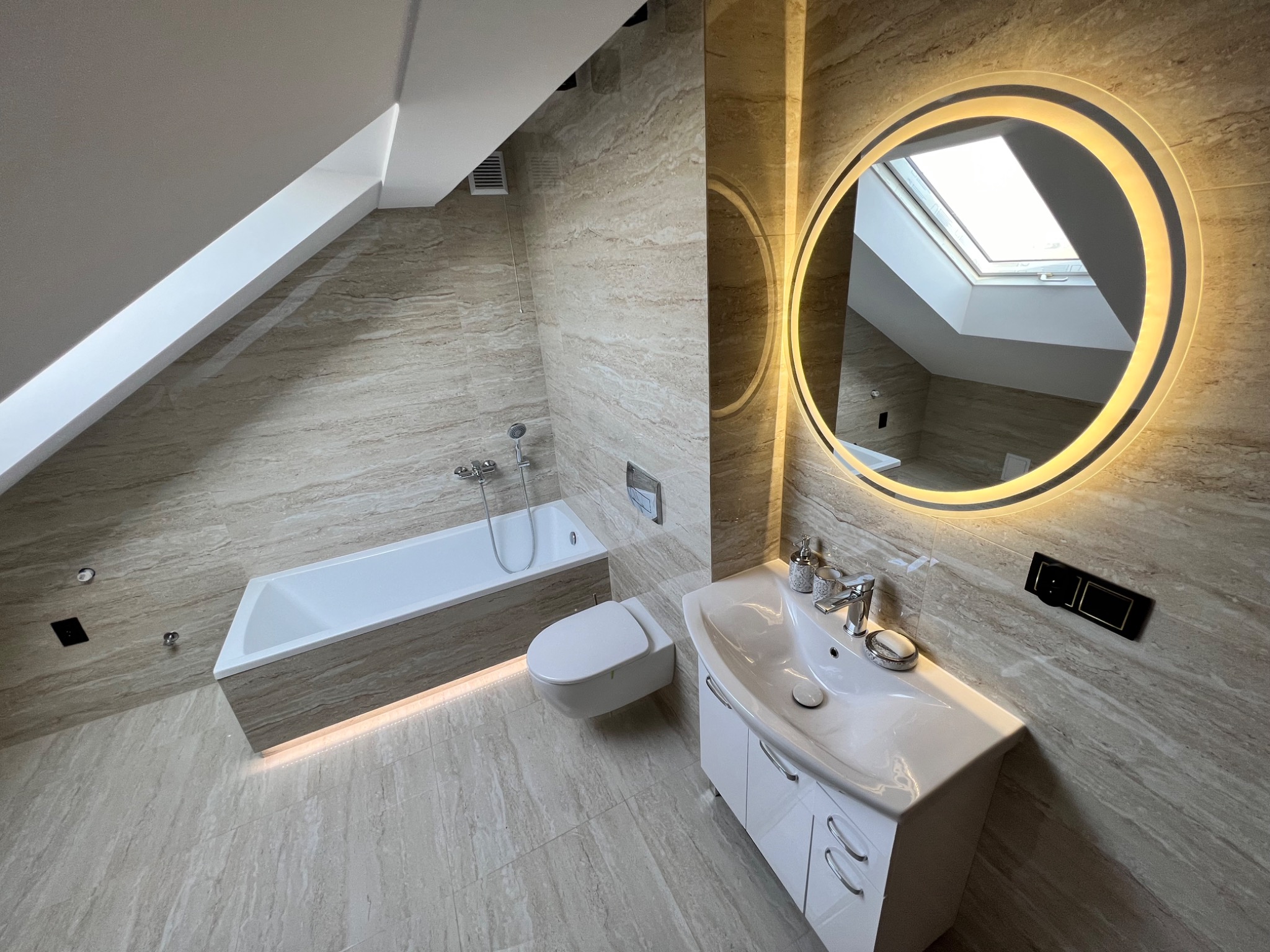 Nowoczesna łazienka na poddaszu z beżowymi płytkami imitującymi marmur, podświetlana wanna, okrągłe lustro z ledowym oświetleniem i widokiem na okno dachowe.