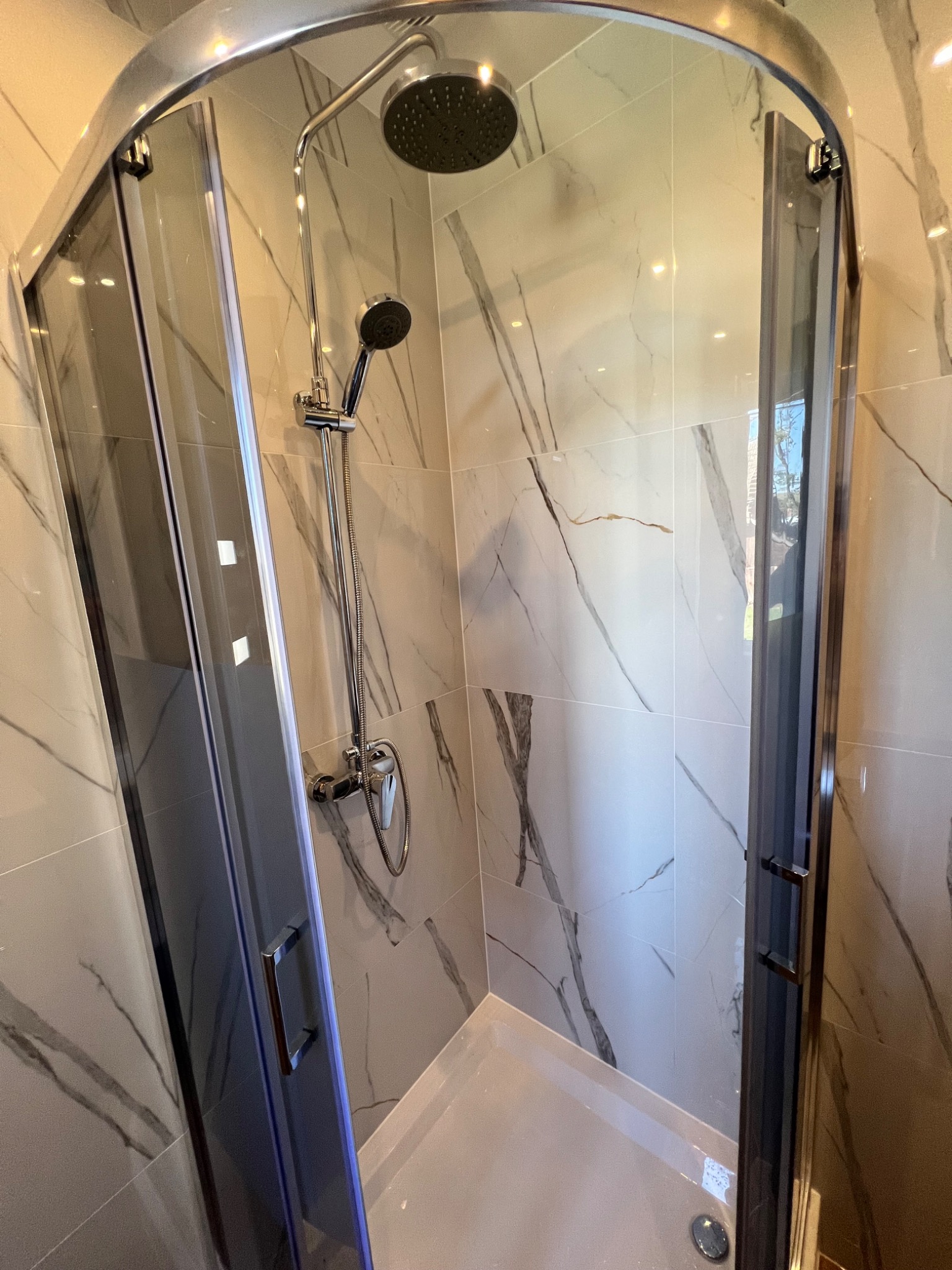 Otwarta kabina prysznicowa z chromowaną armaturą i marmurowymi płytkami na ścianach, widoczny odpływ liniowy w brodziku.