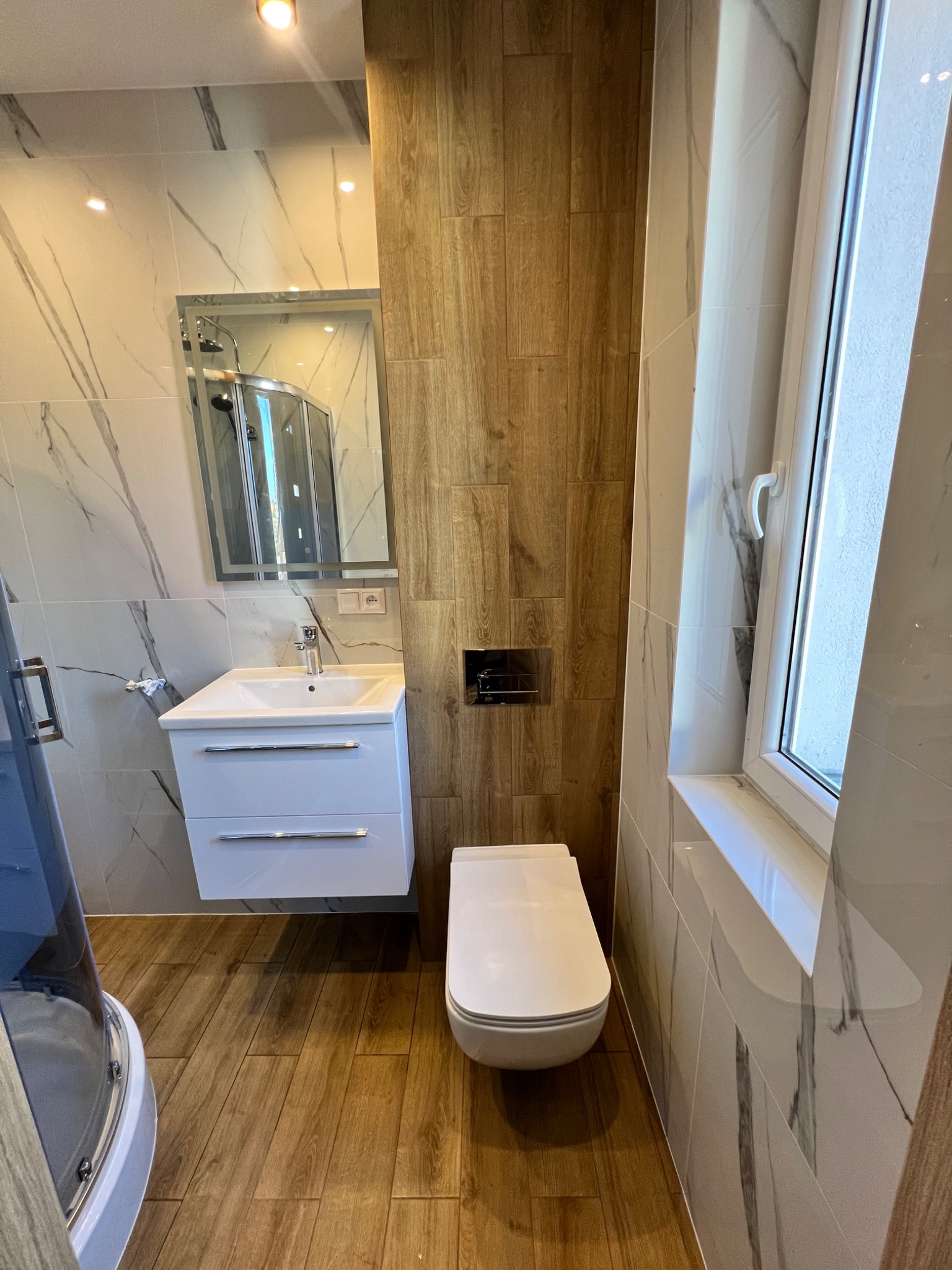 Nowoczesna łazienka z drewnopodobną podłogą i ścianą, białą armaturą, marmurkowymi płytkami oraz kabiną prysznicową widoczną w lustrze.