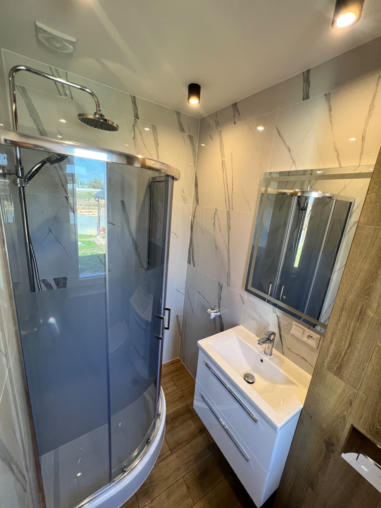 Nowoczesna łazienka z kabiną prysznicową z przyciemnianego szkła, umywalką z szafką w kolorze białym i lustrem w srebrnej ramie, wykończona płytkami imitującymi marmur i drewno.