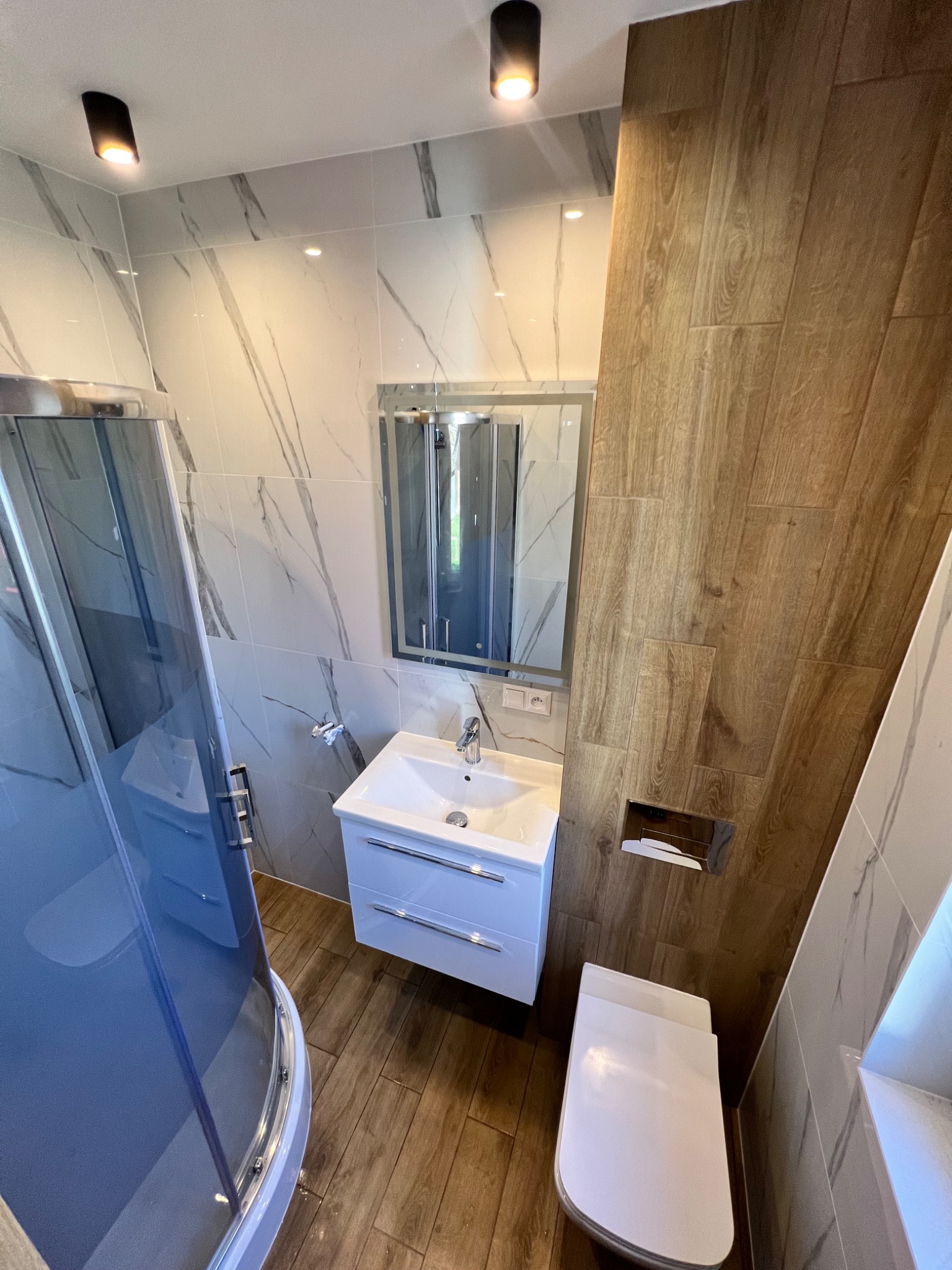 Nowoczesna łazienka z kabiną prysznicową, umywalką z szafką, toaletą i lustrem, wykończona płytkami imitującymi marmur i drewno.