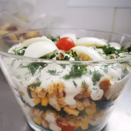 Warstwowa sałatka w szklanej misce: kukurydza, kurczak, pomidory, sos, jajka na twardo i koperek, całość na metalowym blacie.