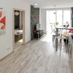 Nowoczesne wnętrze mieszkania: jasna podłoga, ściana z imitacji kamienia, minimalistyczna kuchnia z barkiem i widok na balkon z jadalnią; na ścianie obraz z czerwonymi tulipanami.