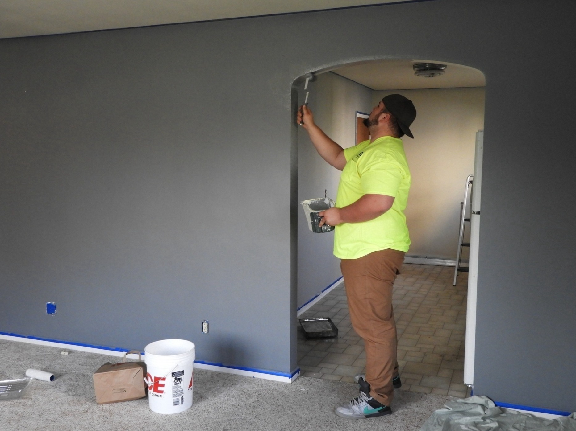 Mężczyzna w neonowej koszulce maluje szarą farbą ścianę w pomieszczeniu, używając małego wałka, widoczne taśmy malarskie, puszka z farbą i akcesoria malarskie leżące na podłodze.