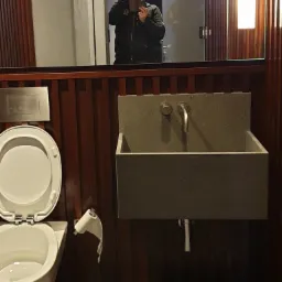 Nowoczesna łazienka z umywalką z betonu architektonicznego, toaletą podwieszaną i widocznym odbiciem osoby robiącej zdjęcie w lustrze.