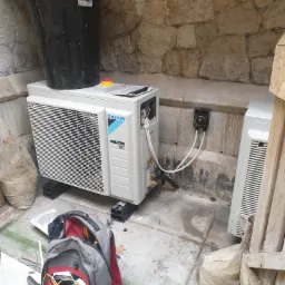 Zewnętrzna jednostka klimatyzacyjna Daikin zamontowana na betonowym podłożu, z widocznymi przewodami i otwartym panelem elektrycznym, obok stoi torba narzędziowa i drugi moduł klimatyzacji na tle...