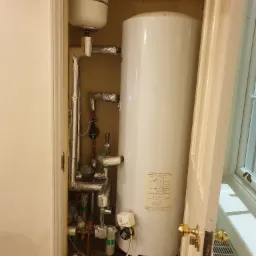 Bojler w szafie z widocznymi rurami, zaworami i elementami instalacji, częściowo zaizolowanymi folią aluminiową, z otwartymi drzwiami szafy i grzejnikiem pod oknem w tle.