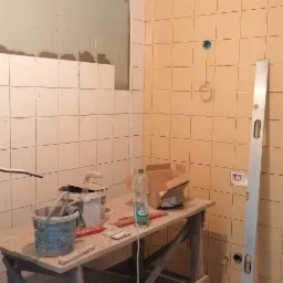 Łazienka w trakcie remontu: częściowo położone żółte płytki, fragment ściany z płyt karton-gips, drewniany stół roboczy z narzędziami, poziomica oparta o ścianę.