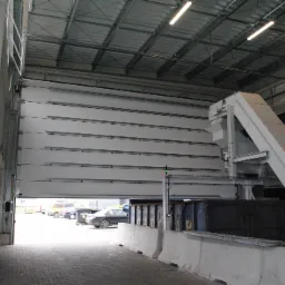 Otwarta segmentowa brama przemysłowa z widocznym mechanizmem podnoszenia, z zewnątrz widoczne zaparkowane samochody i elementy otoczenia.