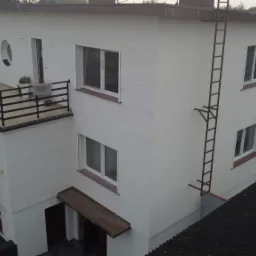 Trzykondygnacyjny budynek z ocieploną, pomalowaną na biało elewacją, zewnętrzną spiralną klatką schodową prowadzącą na balkon z okrągłym oknem i przymocowaną drabiną przeciwpożarową.