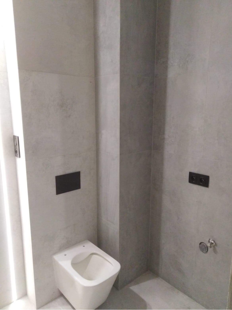 Nowoczesna toaleta podtynkowa w minimalistycznej łazience wyłożonej szarymi płytkami imitującymi beton, z czarnymi elementami armatury i osprzętu elektrycznego.