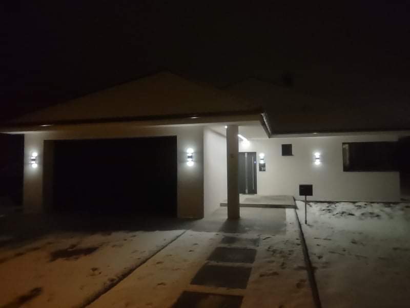 Nowoczesny dom jednorodzinny z garażem, oświetlony zewnętrznymi lampami ściennymi, sfotografowany nocą, po lekkim opadzie śniegu.