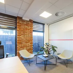 Nowoczesne biuro z ceglaną ścianą, roletami okiennymi, minimalistycznymi krzesłami i roślinami doniczkowymi, oświetlone panelami LED w suficie.