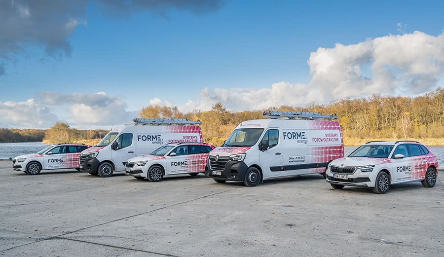 Flota pojazdów firmowych z logo i grafiką paneli słonecznych zaparkowana nad brzegiem jeziora w słoneczny dzień. Na dachach dwóch vanów drabiny.