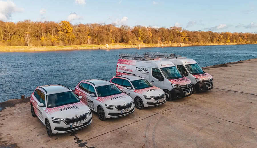 Flota firmowa - dwa samochody osobowe Skoda i dwa busy Renault, wszystkie oklejone logo firmy FORME Energy na tle rzeki i jesiennych drzew.