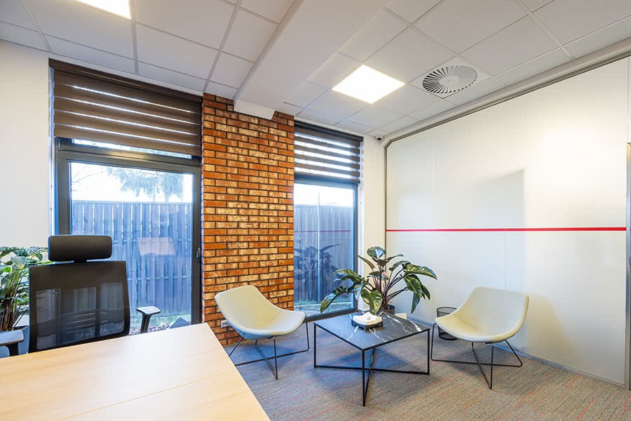 Nowoczesne biuro z ceglaną ścianą, roletami okiennymi, minimalistycznymi krzesłami i roślinami doniczkowymi, oświetlone panelami LED w suficie.