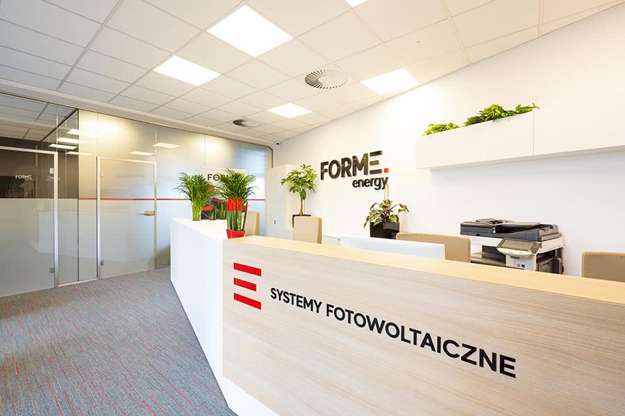 Wnętrze biura firmy z Wrocławia specjalizującej się w systemach fotowoltaicznych, z recepcją z napisem 'Systemy Fotowoltaiczne' i logo firmy 'FORME energy' na ścianie, oraz roślinami doniczkowymi...