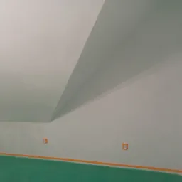 Pokój na poddaszu przygotowany do malowania: podłoga zabezpieczona zieloną folią, listwy przypodłogowe oklejone pomarańczową taśmą, ściany świeżo pomalowane na biało, widoczne gniazdka elektryczne.