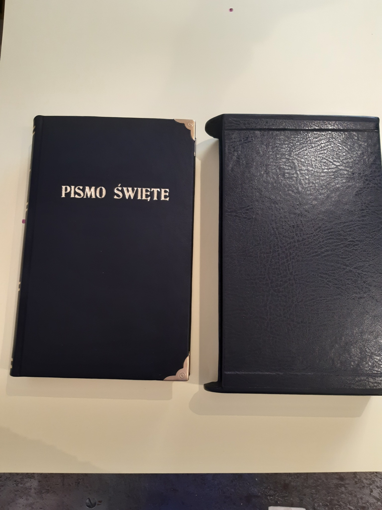 Granatowa oprawa Pisma Świętego z pozłacanymi narożnikami, obok granatowe etui na książkę z fakturą skóry.