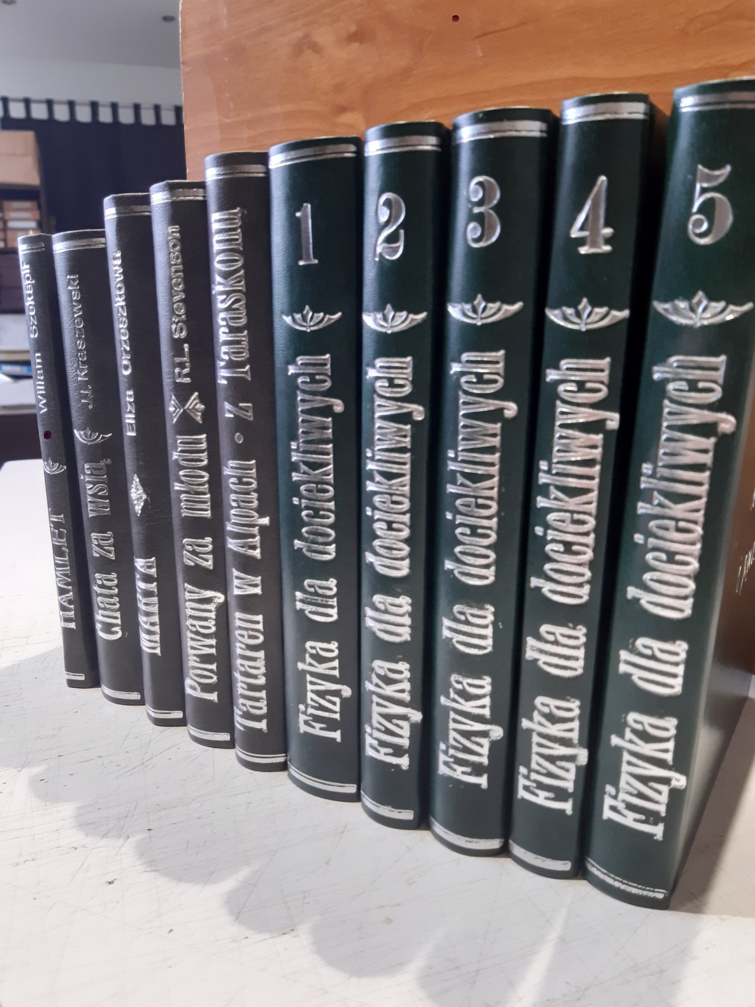 Rząd książek w ciemnych okładkach, od lewej: tom z dramatem Szekspira 'Hamlet', powieść Kraszewskiego 'Chata za wsią', powieść Orzeszkowej 'Marta', powieść Stevensona 'Porwany za młodu' i tomy 1-5...