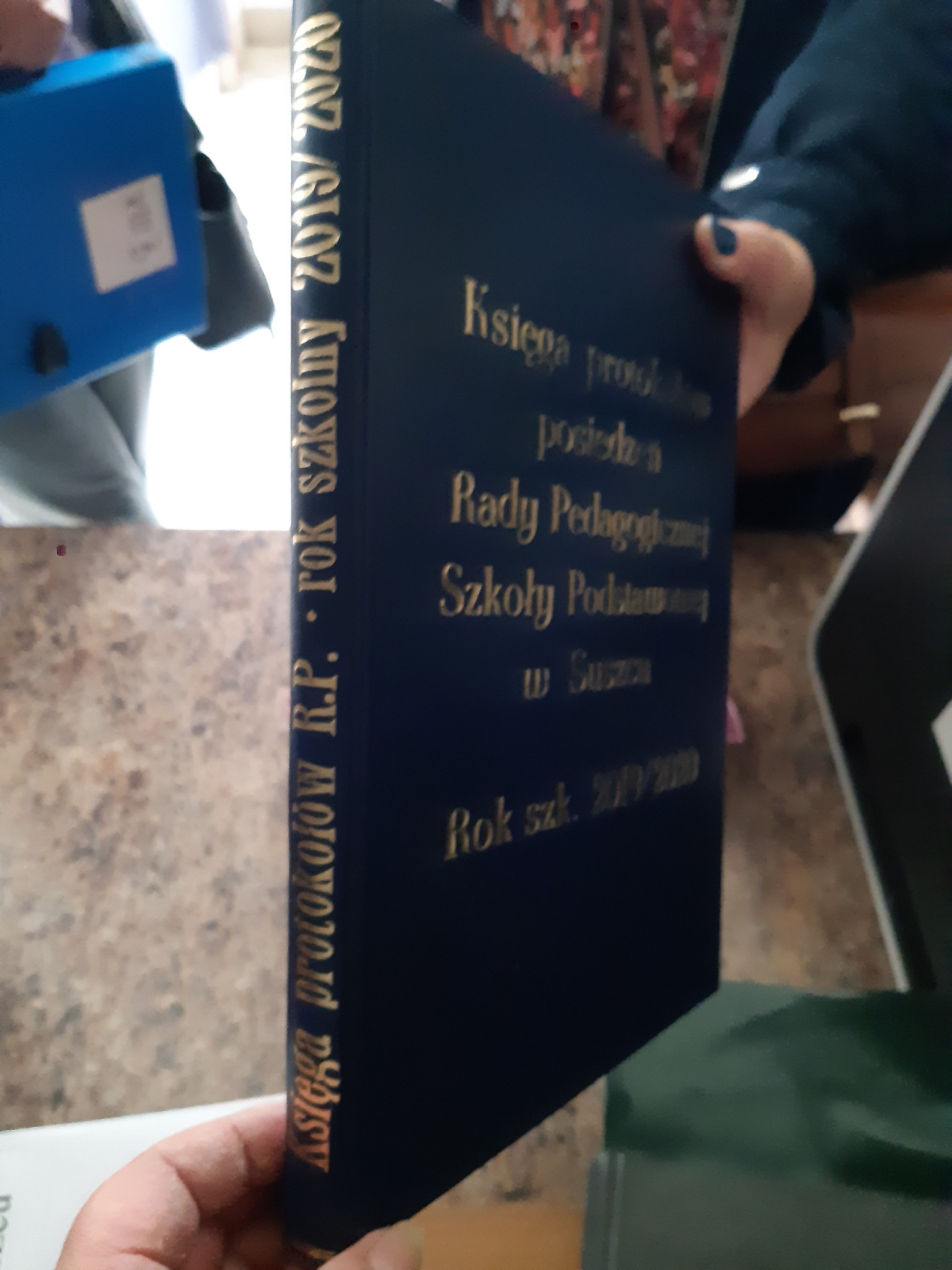 Granatowa, oprawiona księga protokołów Rady Pedagogicznej Szkoły Podstawowej z napisem w kolorze złotym, trzymana w dłoniach.