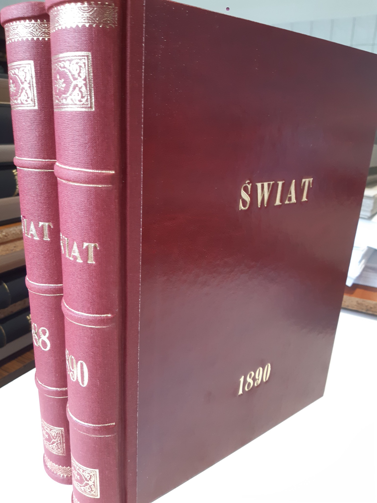 Dwa oprawione w burgundową skórę tomy książki 'Świat' z 1890 roku, zdobione złotymi tłoczeniami na grzbiecie i okładce, ustawione na białym blacie.