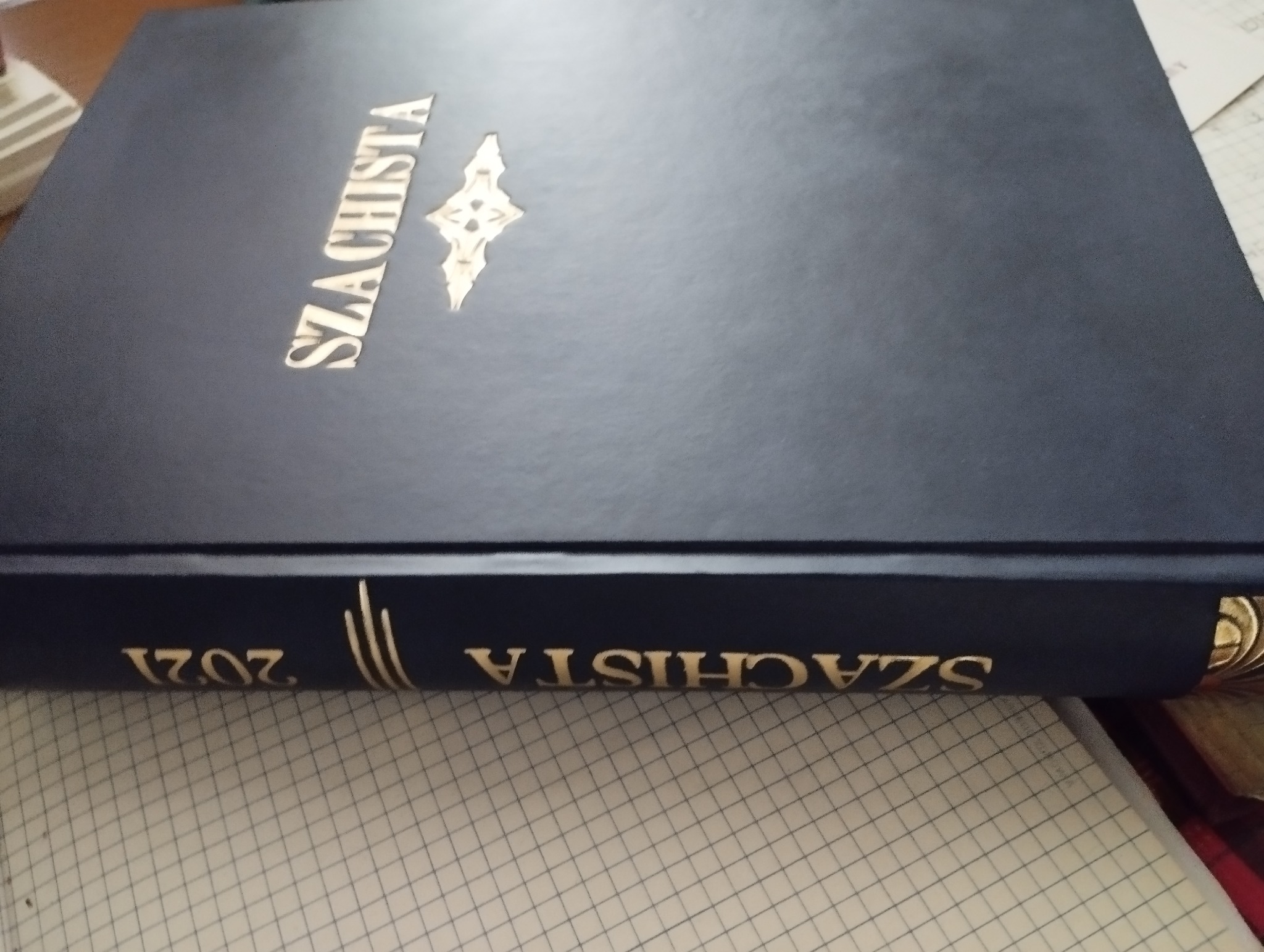 Księga w twardej, granatowej oprawie ze złoconym napisem 'SZACHISTA' i symbolem, leżąca na papierze w kratkę. Na grzbiecie widoczny rok 2021.