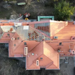 Widok z drona na duży dom z czerwoną dachówką w trakcie remontu dachu; część dachu jest już pokryta nową dachówką, a na pozostałej części widać przygotowane rzędy dachówek.