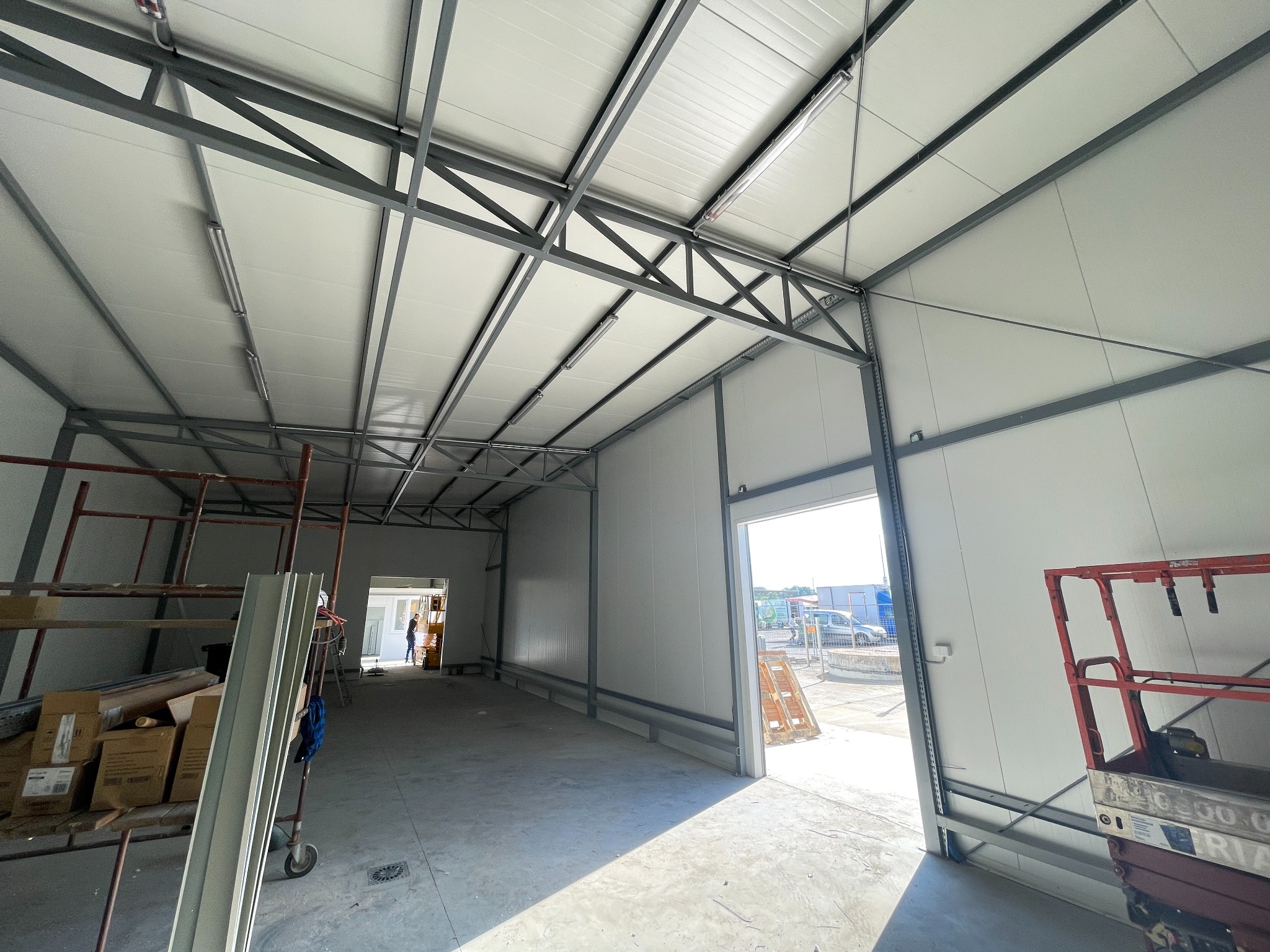Wnętrze hali magazynowej w trakcie budowy, widoczne stalowe konstrukcje nośne, białe panele ścienne i sufitowe, oświetlenie liniowe, oraz częściowo otwarte wrota wjazdowe z widokiem na zewnątrz.