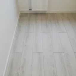 Jasne panele podłogowe z delikatnym wzorem drewna, ułożone w narożniku pokoju, widoczna biała listwa przypodłogowa i grzejnik na ścianie.