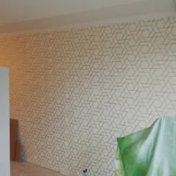 Ściana po tapetowaniu z geometrycznym wzorem, zabezpieczona podłoga i meble folią malarską oraz butelka ze spryskiwaczem w rogu pokoju.